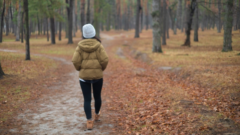 Žena u jakni i kapi hoda šumskom stazom prekrivenom lišćem tokom jeseni